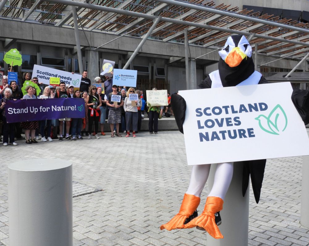 A person in a Puffin costume holds a sign which reads Scotland Loves Nature outside the Scottish Parliament while protesters gather in the background with placards and banners.