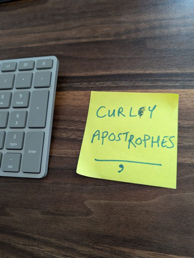 Close up of a yellow post-it note next to a grey keyboard on a dark wood desk. The post-it says in caps 'curley apostrophes' with the 'e' in 'curley' scribbled out.