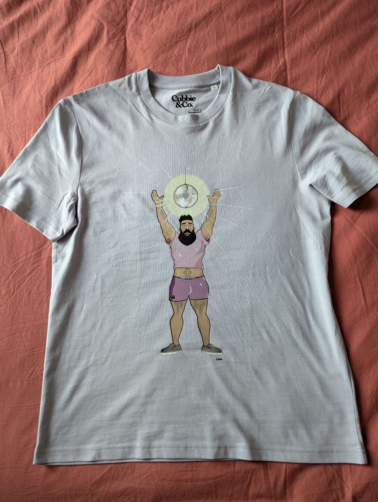 A lilac t-shirt with an illustrated bearded and muscular man in a purple crop top and shorts dancing beneath a glittering disco ball.