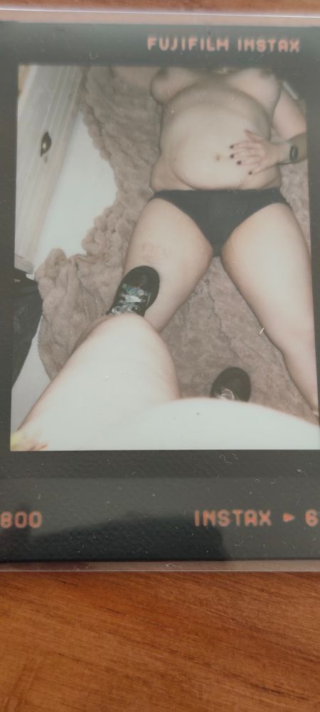 An instant photo showing a point of view of someone standing over a mostly naked woman laying on the floor, with their boot resting on one of her legs 