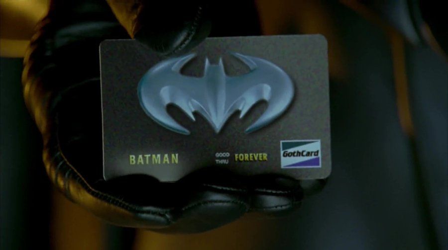 Close up of Batman's hand holding a credit card with the Batman symbol on it. The name "Batman" is inscripted, as well as an expiration date of "good thru forever" and a logo for "Gothcard".