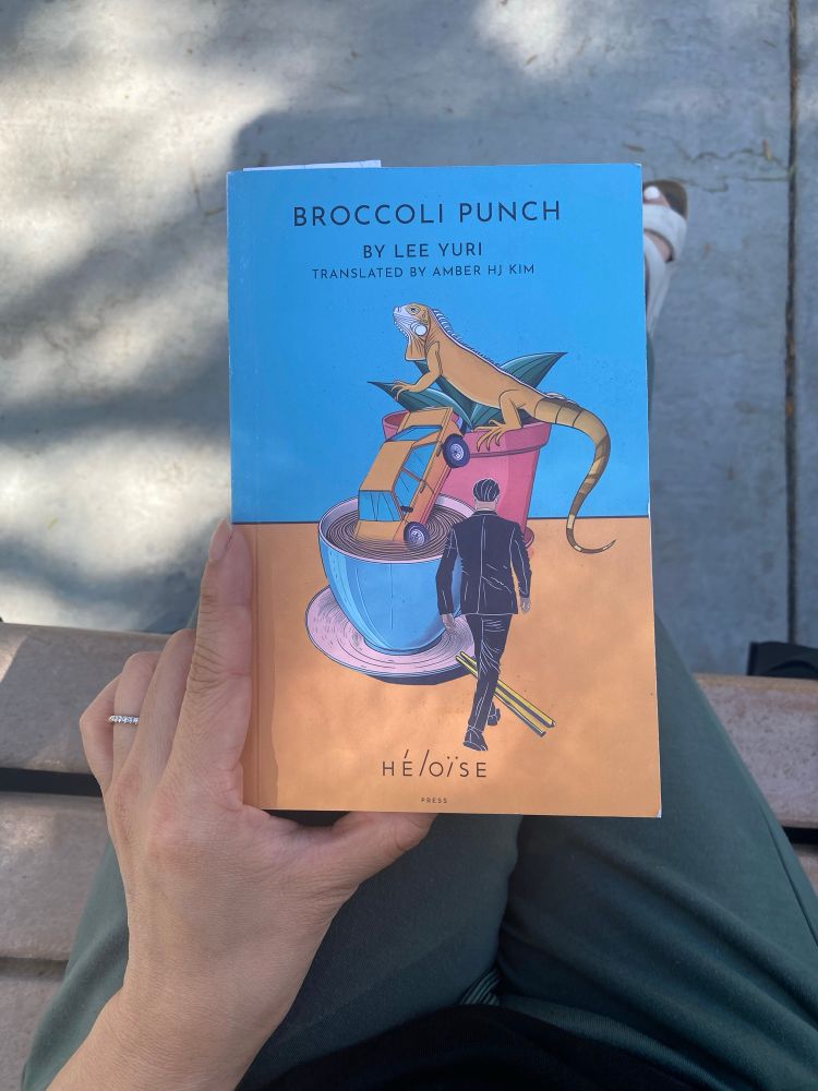 Overhead POV of a book on my lap while sitting on a bench. The book is titled Broccoli Punch by Lee Yuri.