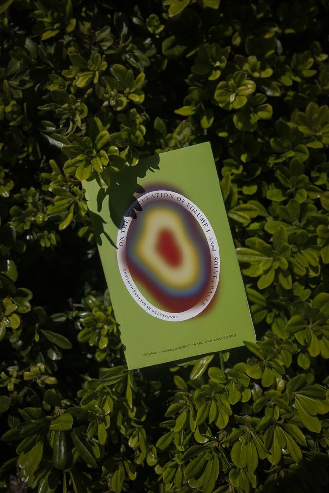 A book with a lime green cover with an oval with different layers of multi-colored rings on the middle. The book is sitting on top of greenery. 
