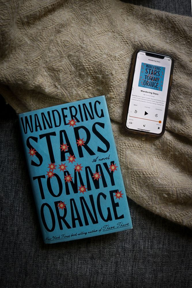 Overhead POV shot of the book Wandering Stars and a smartphone with the audiobook on the screen sitting on top of a beige blanket and gray couch.