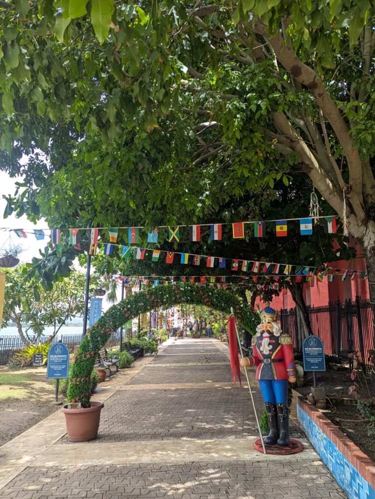 Sidewalk alongside the ocean with small international flags hanging from the trees above. 