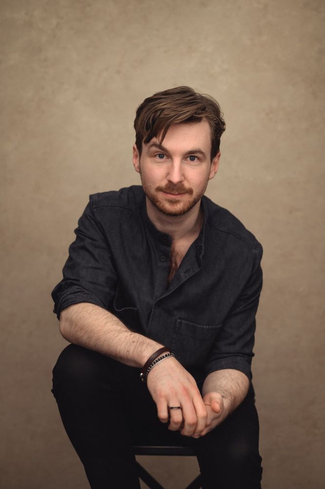 Edward (light skin, wavy brown hair, blue eyes, wearing a collarless dark blue shirt and a mix of black and brown bracelets), sat against a beige backdrop as his elbow leans on one knee, and he smiles at the camera.