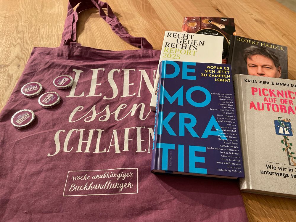 Fünf Bücher, die ich heute gekauft habe:

- Demokratie - Wofür es sich jetzt zu kämpfen lohnt, verschiedene Autoren
- Picknick auf der Autobahn, Katja Diehl und Mario Sixtus
- Recht gegen Rechts Report 2025, verschiedene Autoren 
- Den Bach rauf, Robert Habeck
- Verhängnisvoller Champagner, Tom Hillenbrand

und die lila Stofftasche (Aufdruck in weiß „Lesen
essen
schlafen
Woche unabhängiger Buchhandlungen“)

UND die runden Schokotaler (Aufkleber „Woche unabhängiger Buchhandlungen“) die ich in meiner unabhängigen Buchhandlung dazu geschenkt bekommen habe.

Alles liegt auf einem Holztisch.

