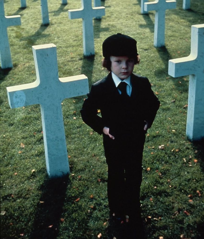 Still from The Omen with Damien standing in a cemetery between crosses.