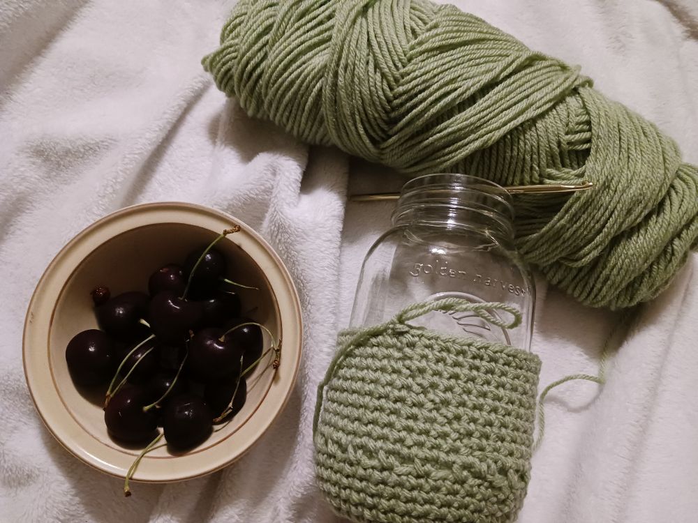 Green yarn, a crochet Mason jar cover in progress, gold crochet hook, bowl of dark cherries, all on a white blanket
