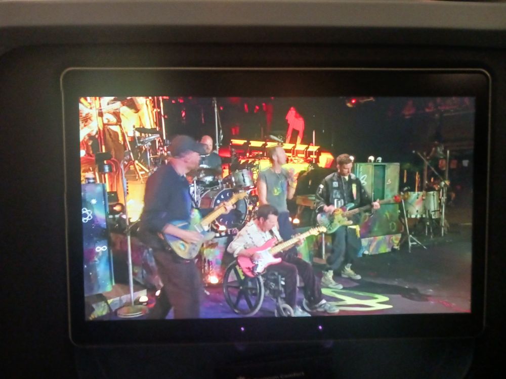 Coldplay and actor Michael J Fox on a stage at Glastonbury music festival. Michael's in the middle sat in a wheelchair playing electric guitar. To the left are Johnny and Will playing electric guitar and drums; to the right are Chris singing and Guy playing electric bass