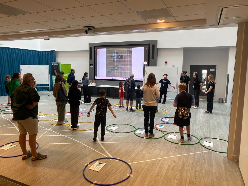 A large room, with several families with small children standing in different hula hoops. The hoops are arranged in a pattern across the floor with lines connecting them.