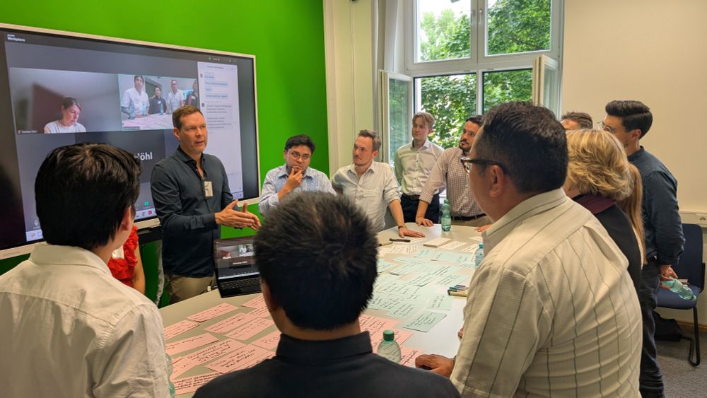 Prof. Johannes Glückler is leading the discussion around the collected topics, projects and contacts. He and the other participants are standing around a large table in the GeoMediaLab.
