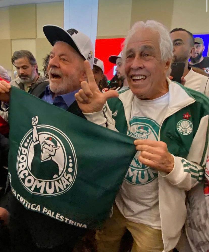 Lula ao lado de um senhor que está com um casaco do palmeiras e fazendo o L. O presidente Lula está de boné, cantando e segurando uma bandeira do porcomunas 