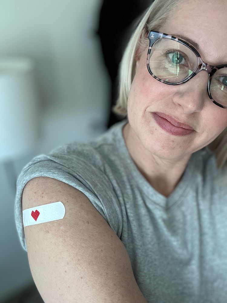 A photo of a woman in a gray t-shirt with a white bandaid with a red heart on it on her arm.