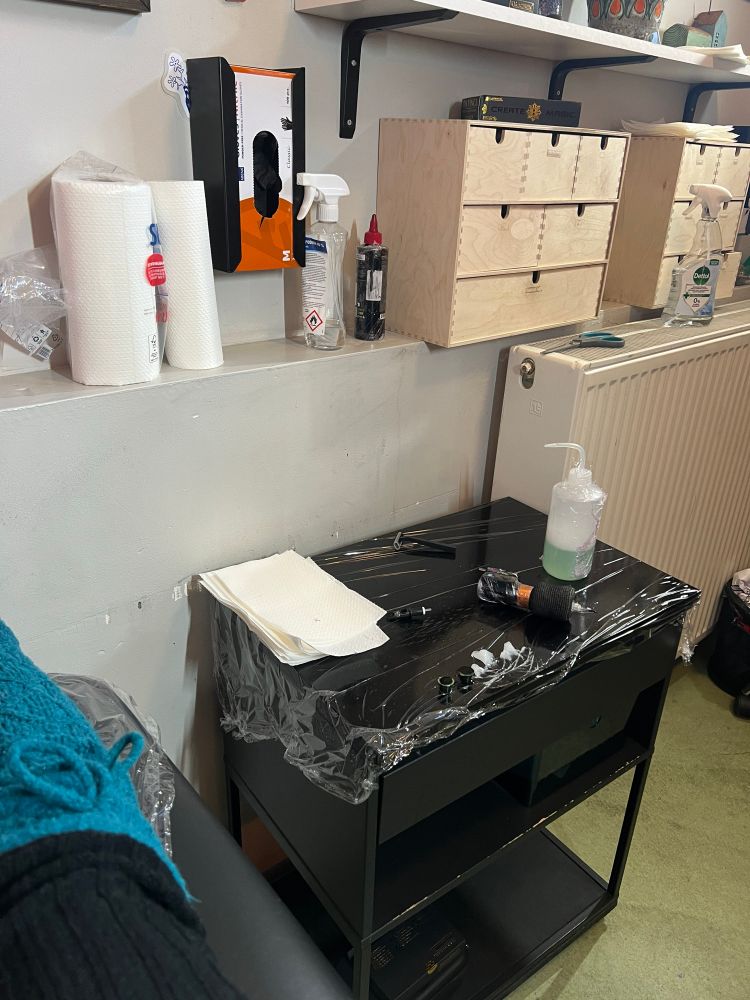 A photo of a set up tattoo workspace with a soap bottle, wipes, a tattoo machine, ink and cream. Behind the trolley, in a shelf, are more hygienic items like, gloves, wipes and tattoo ink. 