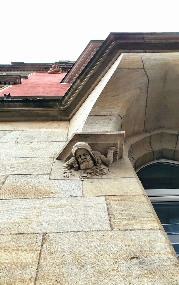 Das Bild zeigt eine steinerne Fassadendekoration an einem historischen Gebäude. Die Perspektive des Fotos ist von unten nach oben gerichtet. Im Vordergrund ist eine Skulptur zu sehen: Ein in Stein gehauener Kopf und Oberkörper eines bärtigen Mannes mit Kapuze, der unter einem Sims hervorschaut, als würde er sich über den Rand beugen. Die Figur scheint aus dem Mauerwerk „herauszukommen“, was ihr einen lebendigen Eindruck verleiht.

Die Fassade selbst besteht aus großen, regelmäßig behauenen Sandsteinblöcken. Oberhalb der Skulptur sind weitere architektonische Details erkennbar, z. B. ein Fensterbogen und Gesimse. Weiter oben auf dem Gebäude ist eine weitere kleinere Figur zu erkennen, die sich mit verschränkten Armen auf eine Kante stützt.


