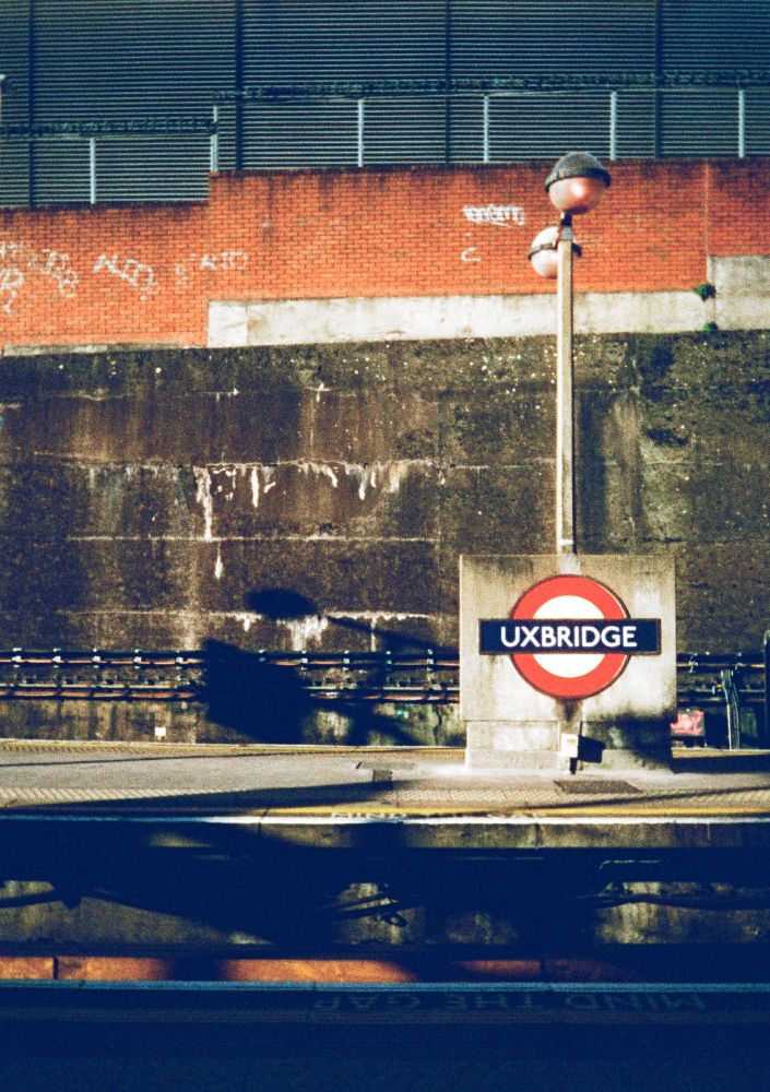High contrast colour film photograph taken in the afternoon sunshine outside on the outdoor portion of the platform at Uxbridge London Underground Station. A roundel bearing the name of the station is on a rectangular concrete block.