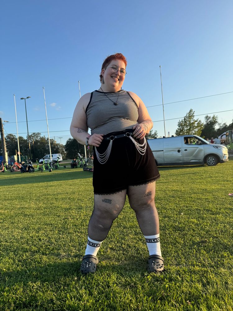 A photo of Michelle, a white fat woman. She is standing outside on a field in sunset lighting. She has short red hair, round glasses, a black and white striped tank top, black velvet shorts, a belt with chains hanging off it, fishnet tights, white tennis socks and black crocs with spikes on. She is looking very happy and confident, smiling broadly at the camera. 
