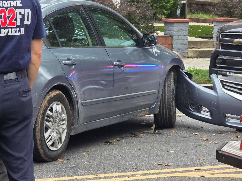The forward part of a light blue Nissan Altima. Its front bumper has been ripped off the car and hangs at an angle on the ground. A firefighter in the foreground has their back to the camera.