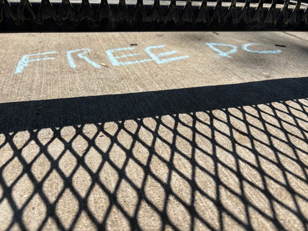Free DC written in chalk under a fence with the shadow of the fence below it