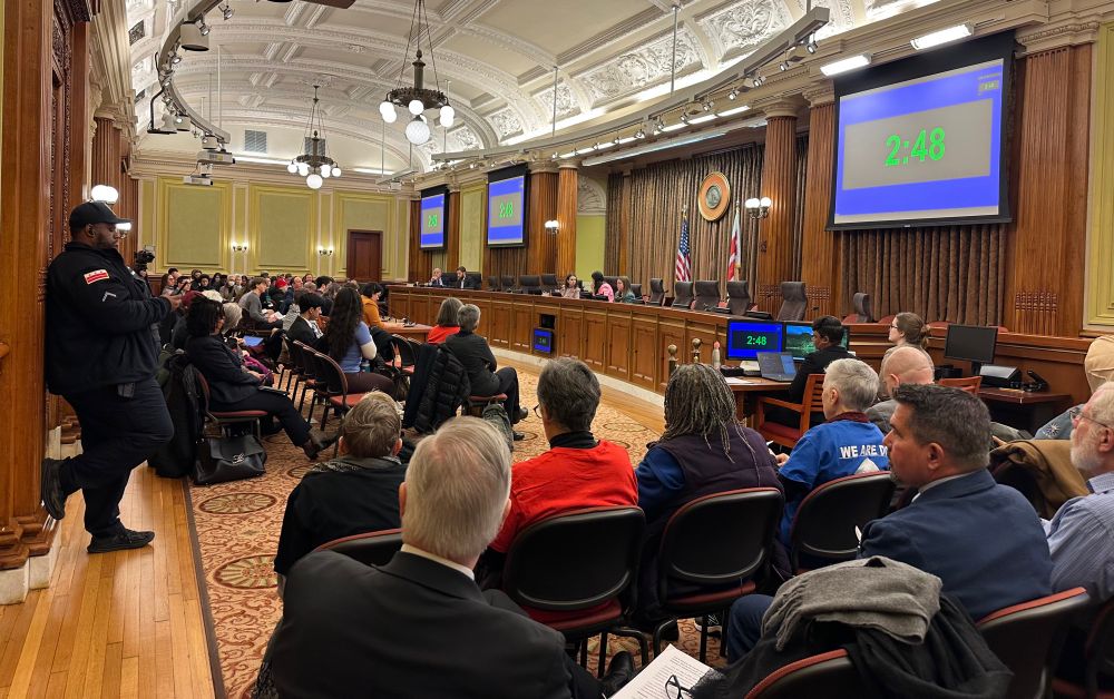 A government hearing room full with people in the audience. Elected official sit at the front at a dais. Large digital screens on the wall show 2 minutes and 48 seconds.