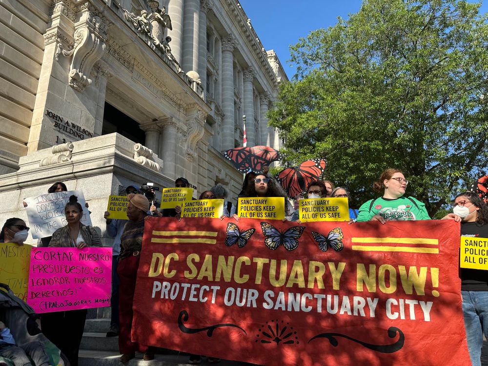Activists hold a banner that reads “DC Sanctuary Now! Protect our sanctuary city.” They stand in front of of the Wilson Building in the warm sunshine.