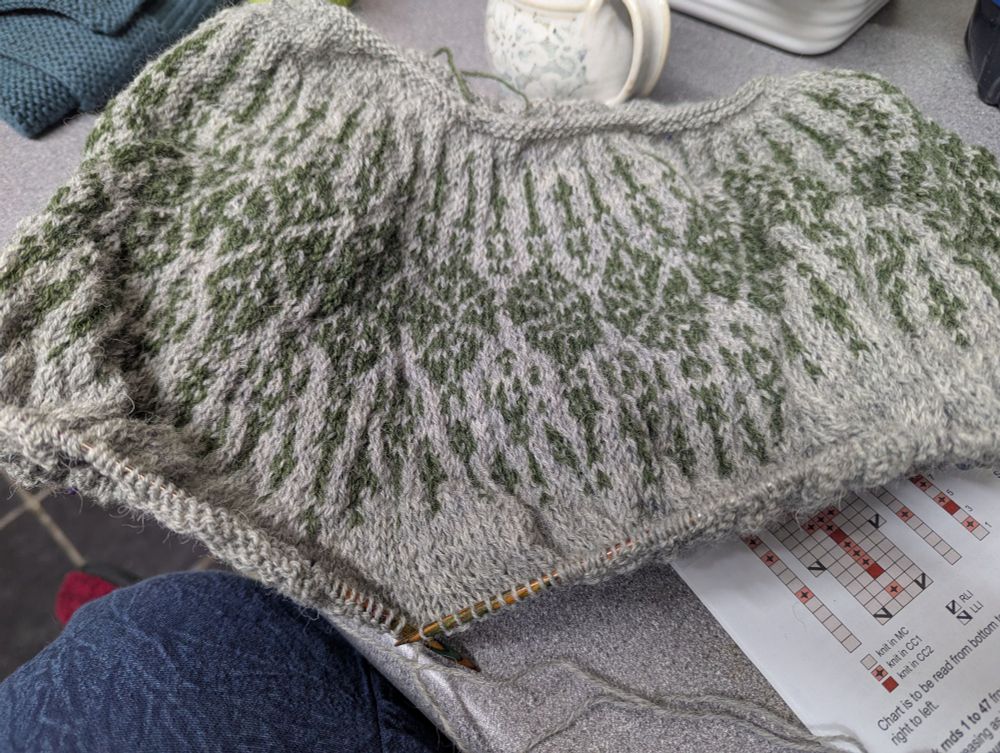 The yoke of a handknitted sweater. Dark sage green hanging bauble motif on a marled mid grey background 