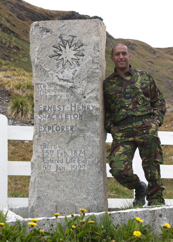 A RAF man wearing camouflage clothing, he is standing next to the grave of Sir Earnest Shackleton on the island of South Georgia.