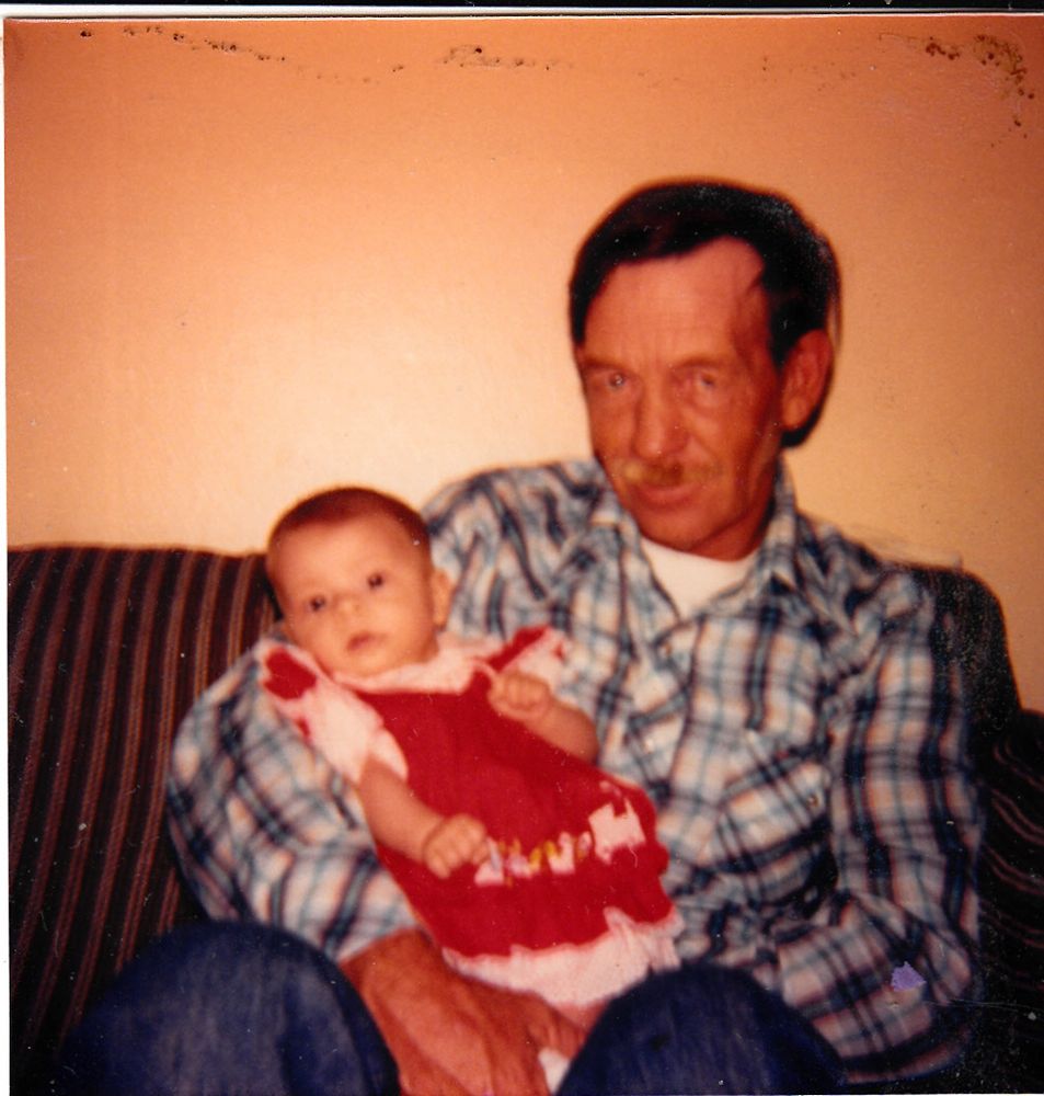Baby Annaleigh at a few months old being held by her dad. She's a little dark hair, dark eyed, light skinned baby wearing a red and white choo choo train dress. Her dad is a mixed Cherokee man in his late 40s, with tan skin, a mustache, and wearing blue jeans and a blue and orange plaid shirt with a long johns undershirt. 