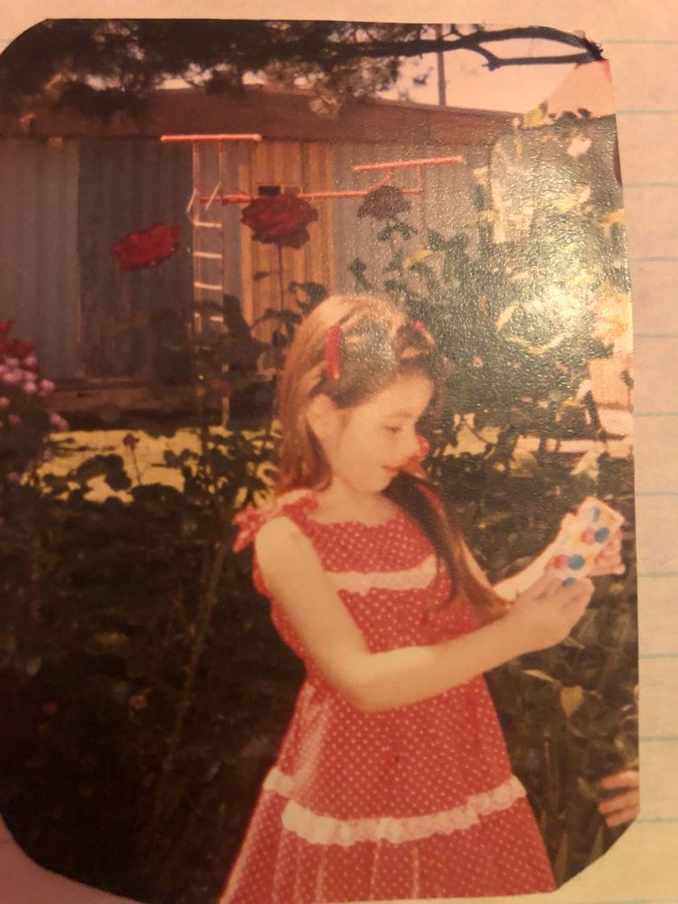 Baby Annaleigh as a five year old. She has long dark hair, dark eyes, light skin. She's wearing a sleeveless red and white polka dot tiered dress and red barrettes in her hair. She's holding and looking at an orange sorbet push up pop, the wrapper is covered in rainbow polka dots. In the background are red and pink roses. 