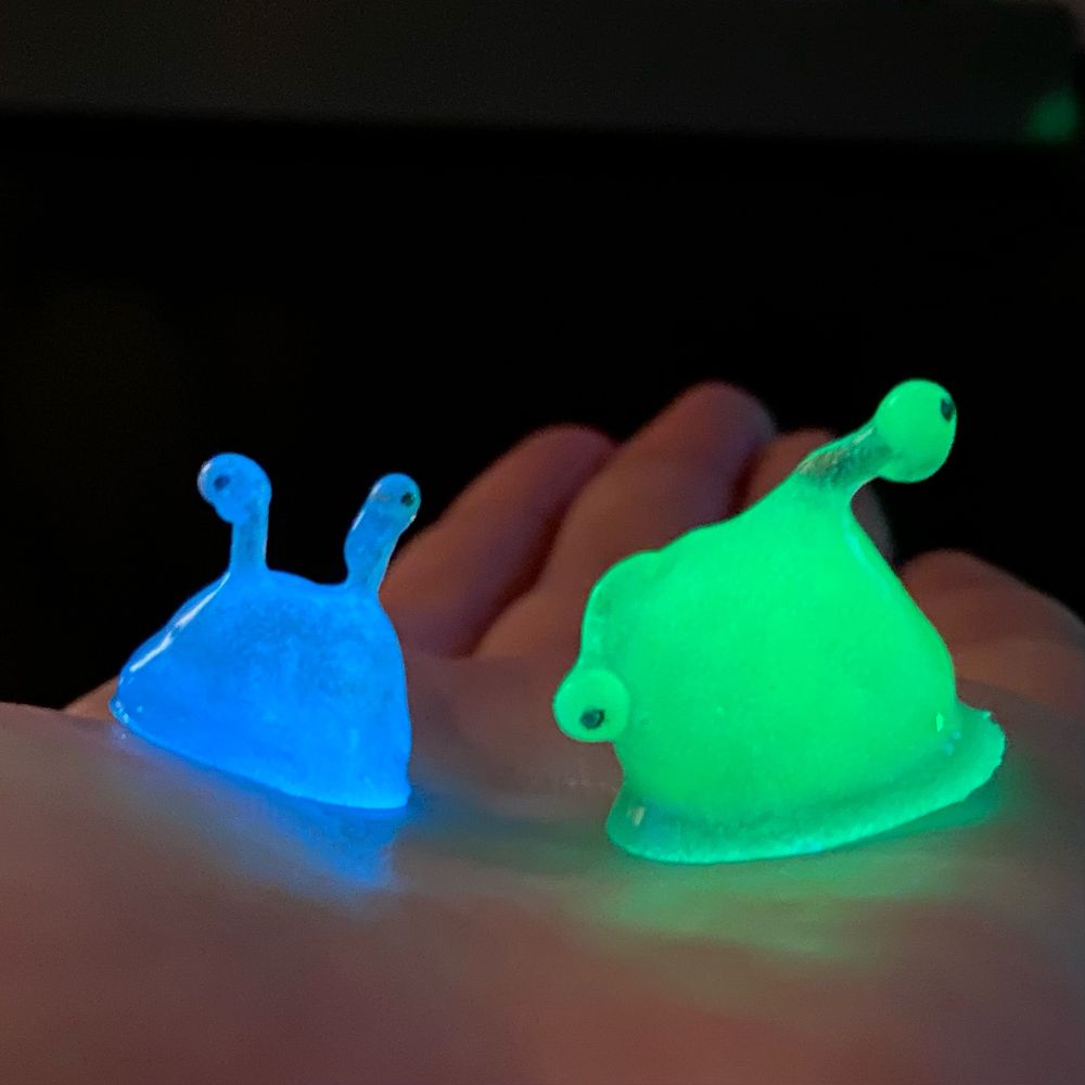 Photo of the little guys held in the palm of a hand in the dark. The blue one glows blue and the green one glows green.