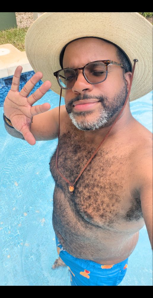 A photo of Kyle, wearing swim trunks with rubber ducks on them, winking and waving at the camera from within the pool
