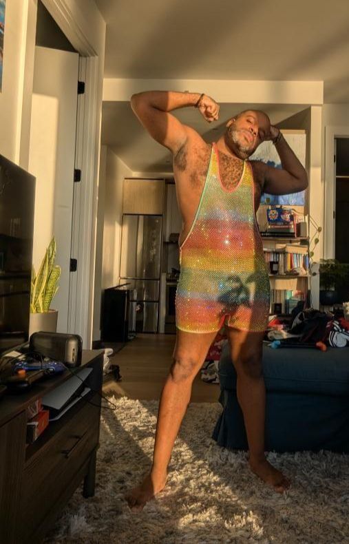 Kyle poses for a selfie in a somewhat untidy living room, wearing a mesh rhinestone rainbow singlet and flexing with his arms raised above his shoulders.