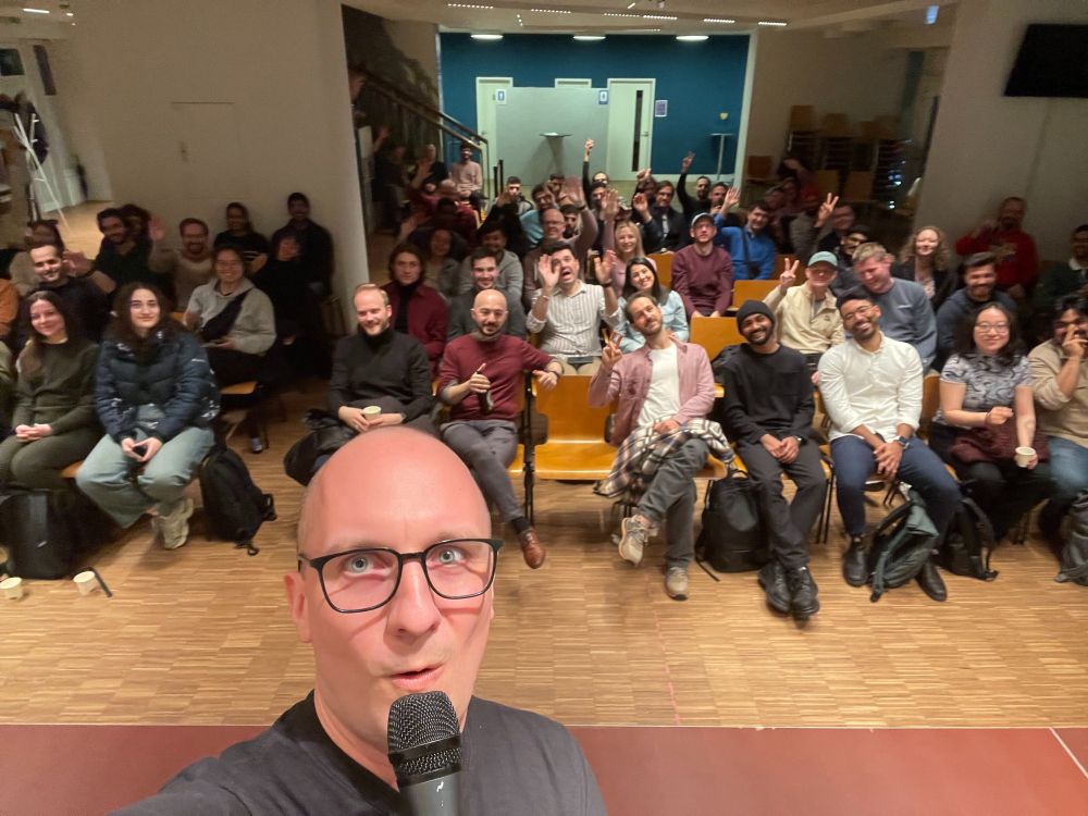 A smiling, bald man named Tim Pietrusky holds a microphone and takes a selfie in front of a large group of people seated in rows. Many in the audience are smiling, waving, or giving thumbs up in a well-lit event space with chairs and an open floor area.