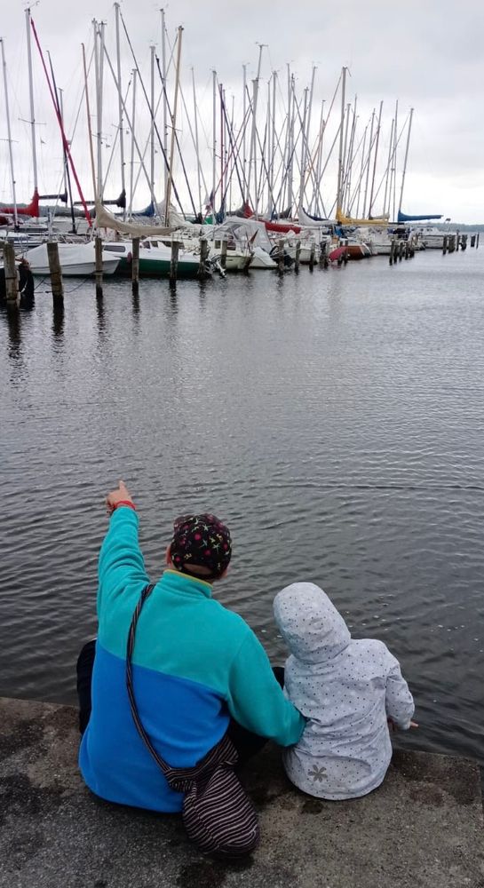 An adult and a child sit on a concrete edge by the water, facing a marina with many sailboats docked. The adult, wearing a turquoise and blue jacket with a patterned hat, points towards the boats, while the child, dressed in a light-colored hooded jacket with star patterns, sits closely beside them. The sky is overcast, and the water is calm, reflecting the masts of the sailboats.