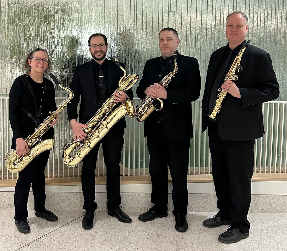 The members of Bacchus Saxophone Quartet stand together in line. Each member of the Quartet is wearing black and holding their instrument.