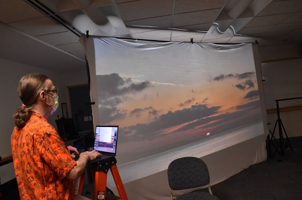 Artist Lorne Covington shows students how their artwork will look projected on a large screen. Lorne stands to the side with his laptop balances on top of a ladder. The projected image is a sunset over the ocean.
