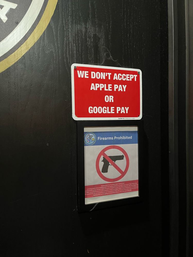 A photo of two signs at the entrance to a store. One says "firearms prohibited" and the other, larger sign says "we don't accept Apple Pay or Google Pay"