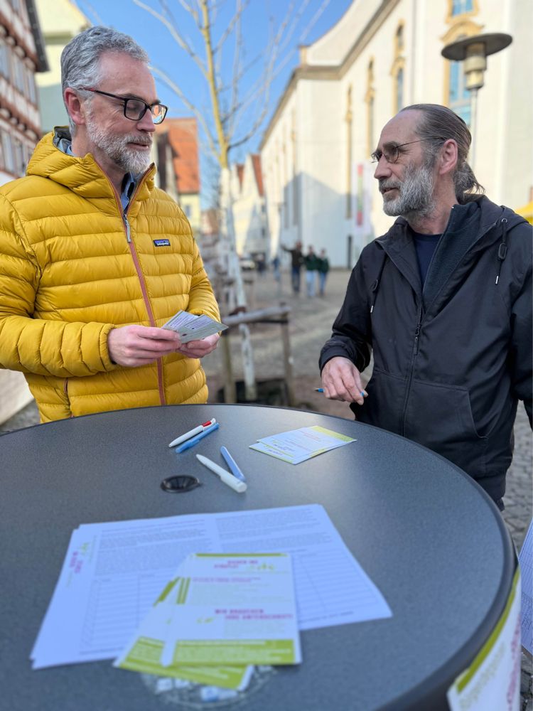 Zwei Menschen stehen an einem Tisch und unterhalten sich. Sonne scheint im Hintergrund