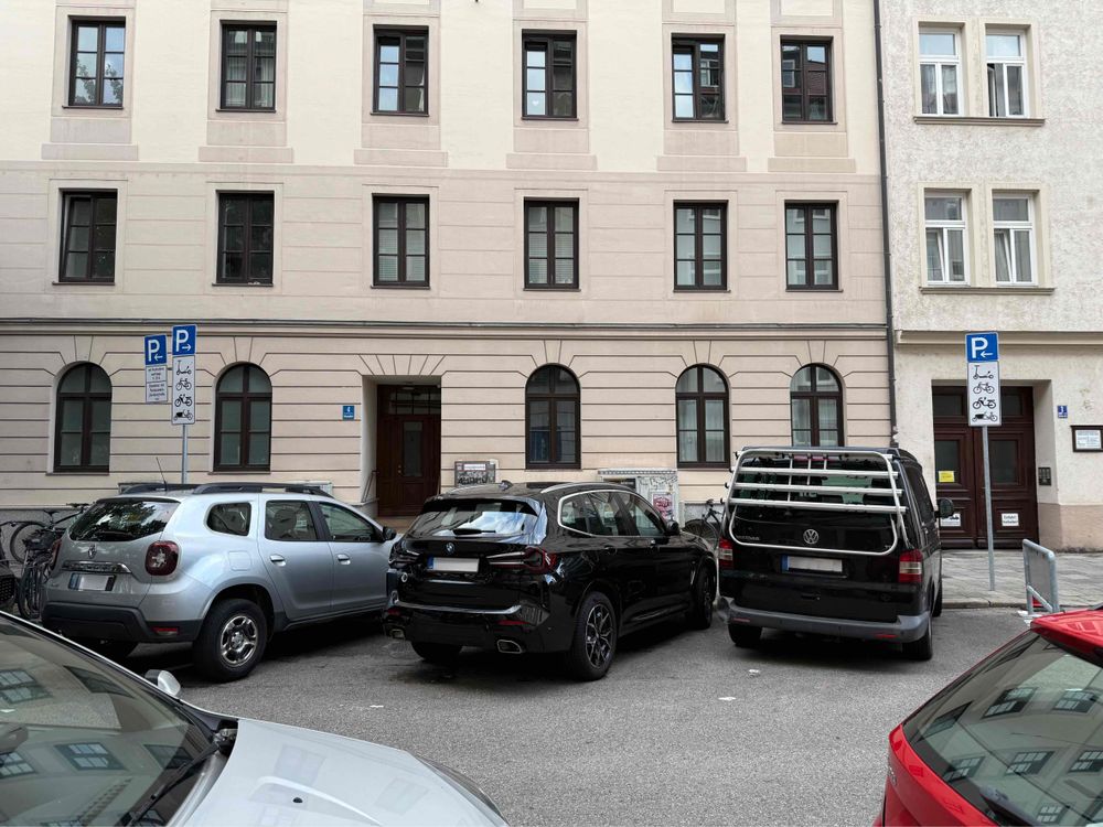 Das Foto zeigt eine Straße vor einem hellen Altbau in einer Stadt. Drei Autos (ein silberner SUV, ein schwarzer PKW und ein schwarzer VW-Bus) parken nebeneinander auf einer Fläche, die durch Verkehrsschilder als Parkplatz für verschiedene Fahrräder ausgewiesen ist. Fahrräder stehen auf dem Gehweg neben den Autos. Im Vordergrund sind die Motorhauben zweier weiterer Autos teilweise zu sehen.

