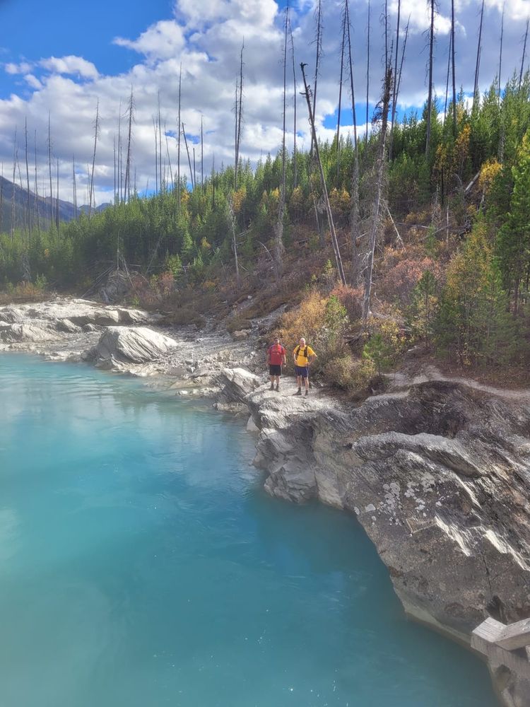 A bright sunny day. Two tiny figures in bright shirts, one in red, the other in yellow, stand on a steep grey limestone riverbank above a pool of an unbelievably auqa marine blue mountain river.
Blue sky and scattered white clouds. Tall dead trees stand sparcely over a new young forest of dense pine trees with patches of trees and shrubs showing yellow fall colors. The valley stretches into the distance. 