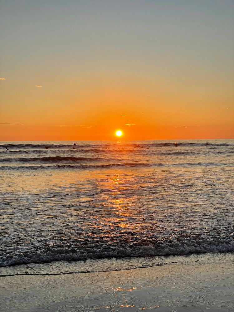 The sun sets over the ocean as orange light shimmers on the surface and waves approach the sand.