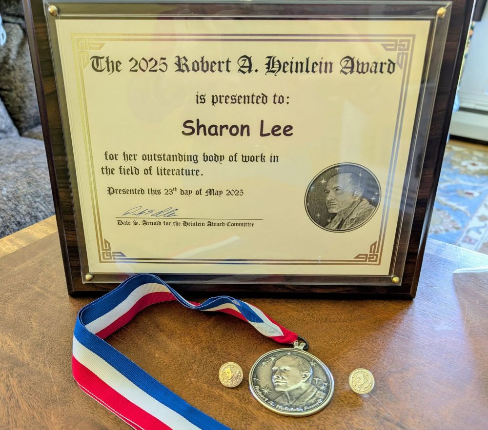A silver medallion on a red, white, and blue ribbon, flanked by two silver lapel pins. Behind a plaque that reads: The 2025 Robert A. Heinlein Award is presented to Sharon Lee for her outstanding body of work in the field of literature.  Presented this 23rd day of May 2025.  Signature, Dale S. Arnold for the Heinlein Award Committee