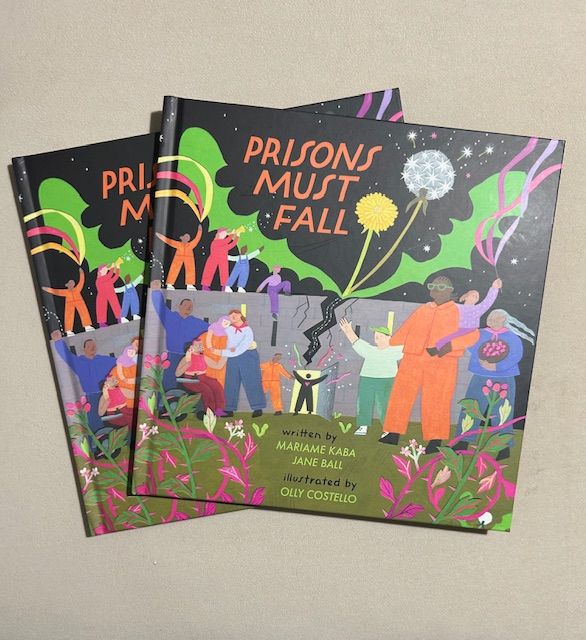 Cover of a book titled "Prisons Must Fall," showing a colorful illustration with diverse people holding hands, flowers, and breaking chains. The book is written by Mariame Kaba and Jane Ball, illustrated by Olly Costello. Two copies are shown.