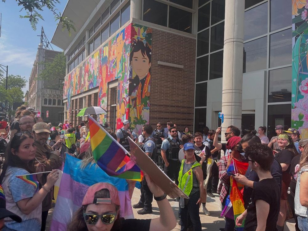 A crowd of people holding Pride flags gathers outside a mural-covered building on a sunny day; some police officers and safety vests are visible among the attendees.