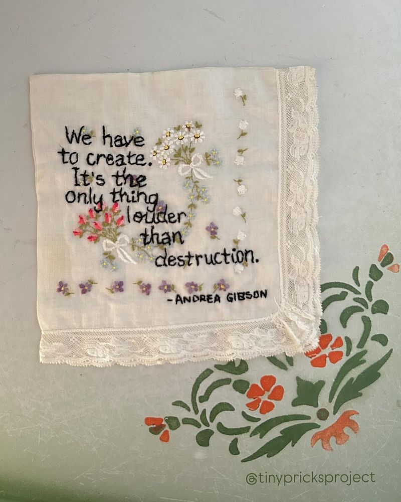 A white handkerchief with lace trim features embroidered flowers and a quote: “We have to create. It’s the only thing louder than destruction. – Andrea Gibson.” The handkerchief sits on a surface with a green and orange floral design. @tinypricksproject