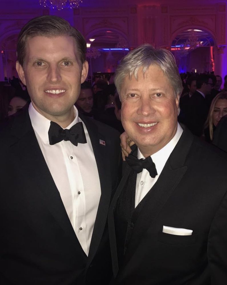 Eric Trump and Morris in formal black tuxedos, smiling at a glamorous event with elegant decor and dim lighting.