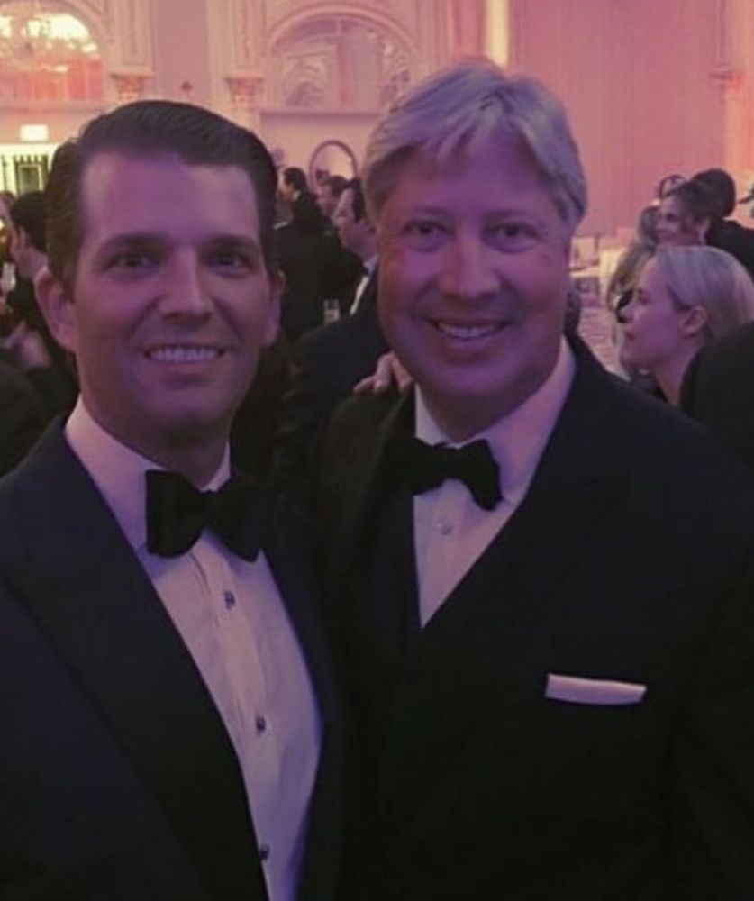 Don Trump jr and Morris in formal black tuxedos stand close together, smiling at a glamorous event with a pink-hued background and well-dressed guests.