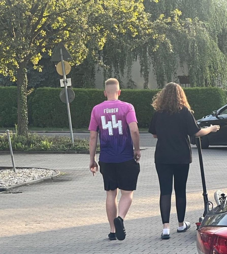 Rückansicht eines jungen Mannes, der ein auffälliges Trikot mit der Aufschrift „FÜHRER“ und der Nummer „44“ trägt. Die Zahl ist in einem Design gestaltet, das an SS-Runen erinnert. Er läuft neben einer jungen Frau auf einem Parkplatz.