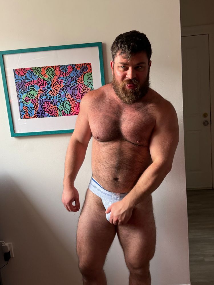 hairy guy in a jock posing while grabbing his crotch, sunlight illuminating one eye